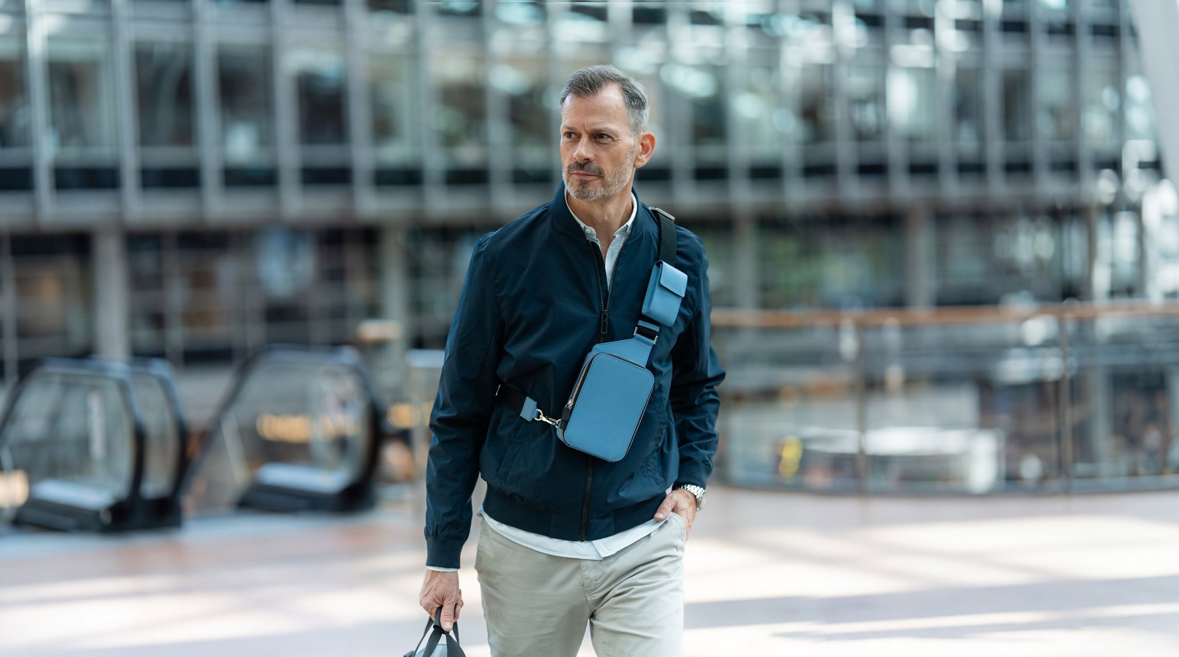 Man walking through a glass-fronted lobby wearing The Verso sling bag, holding a tote bag.